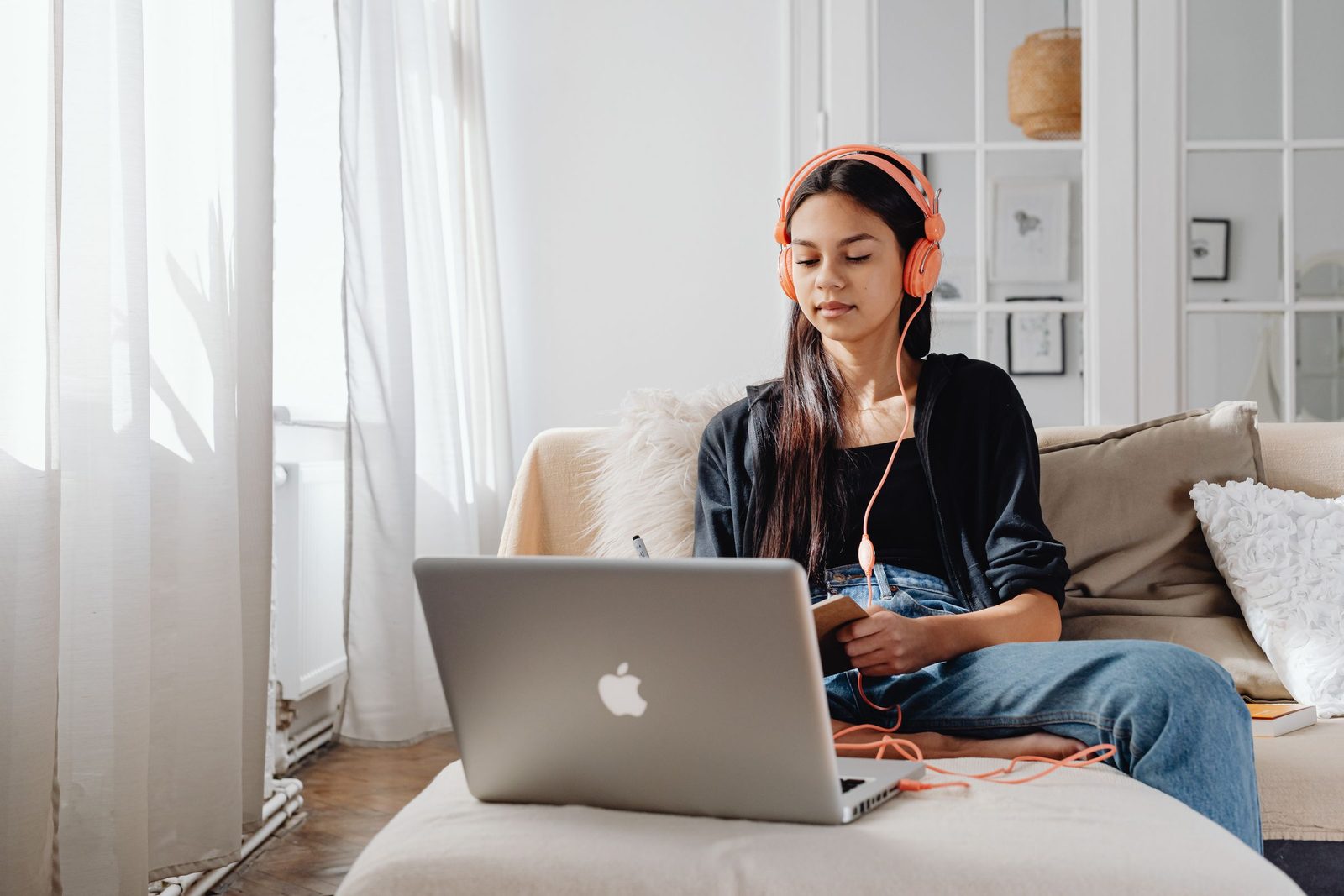 A teenager wearing headphones, sitting on a sofa with a laptop and a notebook